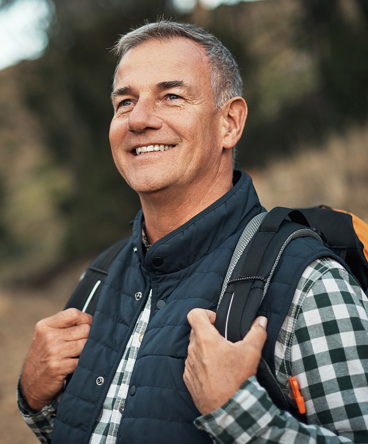Smiling man outdoors with backpack and vest.